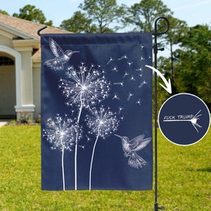 May include: A navy blue garden flag with white dandelion illustrations and two hummingbirds. The flag has the text "FUCK TRUMP" in a circle. The flag is hanging on a black metal pole in a garden.