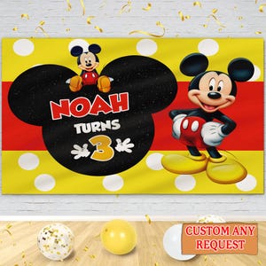 May include: A yellow and red banner featuring Mickey Mouse. The banner has white polka dots and the text "NOAH TURNS 3". Mickey Mouse is depicted in his classic outfit. The banner is likely for a birthday party.