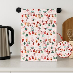 May include: A white Christmas-themed kitchen towel with a repeating pattern of string lights, reindeer, candy canes, and snowflakes. The lights spell out a message. The towel is hanging on a black towel bar.