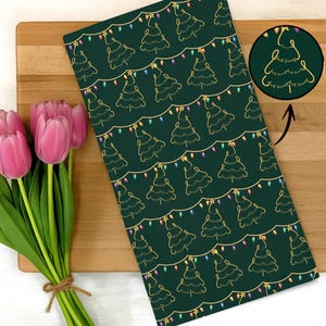 Peut inclure: Un torchon de cuisine vert sur le thème de Noël avec un motif répété d'arbres de Noël dorés et de guirlandes lumineuses. Un petit encart circulaire montre un motif d'arbre avec le texte "F*ck Trump". Des tulipes roses sont à gauche.