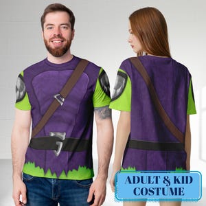 May include: Purple and green costume t-shirts with a brown strap and black belt design. The t-shirts have a jagged green hemline and a graphic on the sleeves. The text "ADULT & KID COSTUME" is displayed on a blue rectangle.