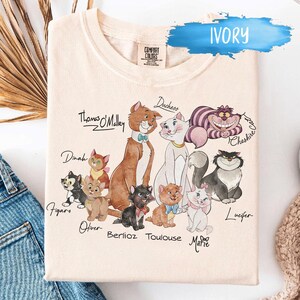 K&ouml;nnte beinhalten: Elfenbeinfarbenes Sweatshirt mit einer Grafik verschiedener Zeichentrickkatzen, darunter Thomas O'Malley, Duchess und Cheshire Cat. Die Namen der Katzen stehen unter jeder Figur. Das Sweatshirt hat ein Comfort Colors-Etikett.