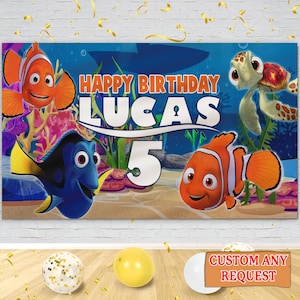 May include: A birthday banner with characters from "Finding Nemo," featuring the text "Happy Birthday Lucas 5." The design includes cartoon fish and underwater scenes. Balloons and confetti are visible in the foreground.