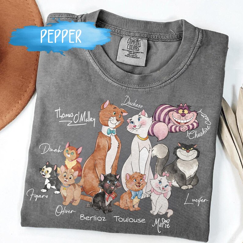K&ouml;nnte beinhalten: Ein graues Comfort Colors T-Shirt mit einer farbenfrohen Illustration verschiedener Zeichentrickkatzen, darunter Thomas O'Malley, Duchess und Cheshire Cat. Die Namen der Katzen sind unter jeder Figur geschrieben. Das Wort "Pepper" steht oben links.