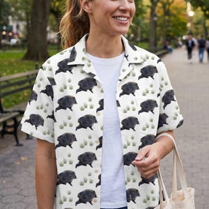 May include: A white short-sleeved button-down shirt featuring an all-over repeating pattern of dark gray wild boars and small green tufts of grass. The shirt has a classic collar, a front button placket, and a single chest pocket on the left side.