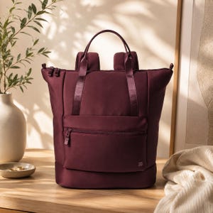 May include: A deep burgundy backpack featuring a structured rectangular silhouette, a front zippered pocket, two top carry handles, and adjustable shoulder straps. The bag includes a small logo detail on the lower right corner of the front pocket.