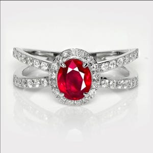 May include: A silver-toned ring featuring a central oval-cut deep red ruby gemstone surrounded by a halo of small white diamonds. The band is split into two crossing sections, each encrusted with a row of small white diamonds along the surface.