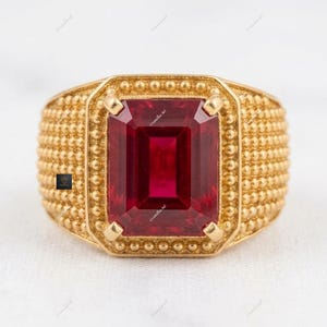 May include: A gold ring with a large, rectangular, deep red gemstone. The ring has a textured band with a dotted pattern and a square setting around the gemstone. The ring is set against a white background.