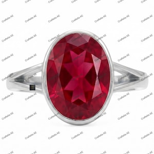 May include: A silver-toned ring featuring a large, oval-cut, deep red gemstone set in a smooth bezel. The band has a split-shank design that tapers toward the sides. The image includes a small logo with the text Craftella AE.