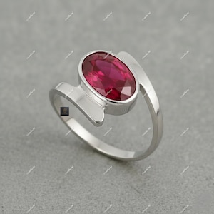 May include: A silver ring featuring a large, oval-cut ruby gemstone. The ruby is a deep red color and is set in a bezel setting. The ring has a modern, minimalist design with a smooth, polished finish.
