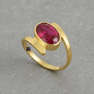 May include: A gold ring with an oval, deep red gemstone. The ring's band has a distinctive, flowing design, with the gemstone set in a bezel. The ring is displayed on a grey surface, highlighting its polished finish.