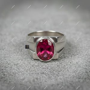 May include: A silver ring featuring a large, oval-cut, deep red gemstone. The ring has a wide band with a rectangular design on the sides, and the gemstone is set in a bezel setting. The ring is displayed on a grey surface.