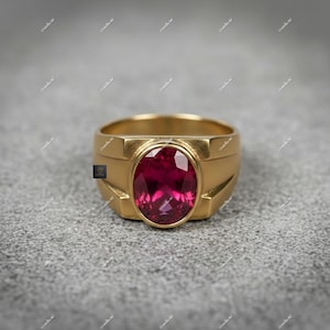 May include: A gold ring featuring a large, oval-cut, deep red gemstone. The ring has a rectangular band with a raised setting. The gemstone is the focal point, reflecting a vibrant colour. The ring's design is classic and bold.
