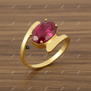 Könnte beinhalten: Ein goldener Ring mit einem großen, oval geschliffenen Rubin. Der Rubin hat eine tiefe, lebendige rote Farbe. Der Ring hat ein einzigartiges, fließendes Design. Der Ring befindet sich vor einem warmen Holzuntergrund.