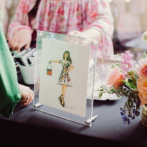 May include: A framed illustration of a woman wearing a floral dress and holding a handbag. The illustration is on a white background and is framed in a clear acrylic frame.