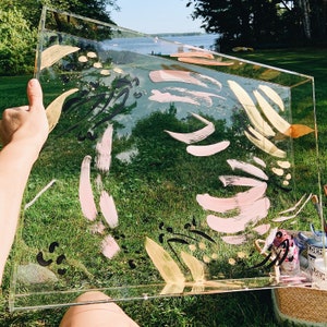May include: A clear acrylic tray with a painted design featuring abstract shapes in black, pink, and gold. The tray is sitting on a grassy surface.