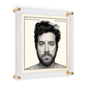 May include: A black and white photo of a man with a beard. The photo is framed in a clear acrylic frame with gold metal corners. The title "Burden of Proof" and the artist's name "Bob Schneider" are printed in red at the top of the photo.
