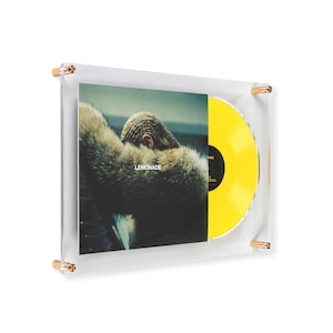 May include: A clear acrylic frame displays a vinyl record with a yellow disc and album cover. The album cover features a person with a fur coat and the word "LEMONADE". The frame has gold-colored hardware.