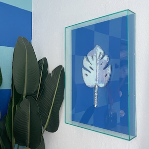 May include: A framed artwork featuring a blue and white mosaic leaf design on a blue background. The frame is made of glass and has a silver finish.