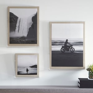 May include: Three framed black and white photographs on a white wall. The top photo shows a waterfall in a mountainous landscape. The middle photo shows a person riding a motorcycle on a beach. The bottom photo shows a person standing on a beach.