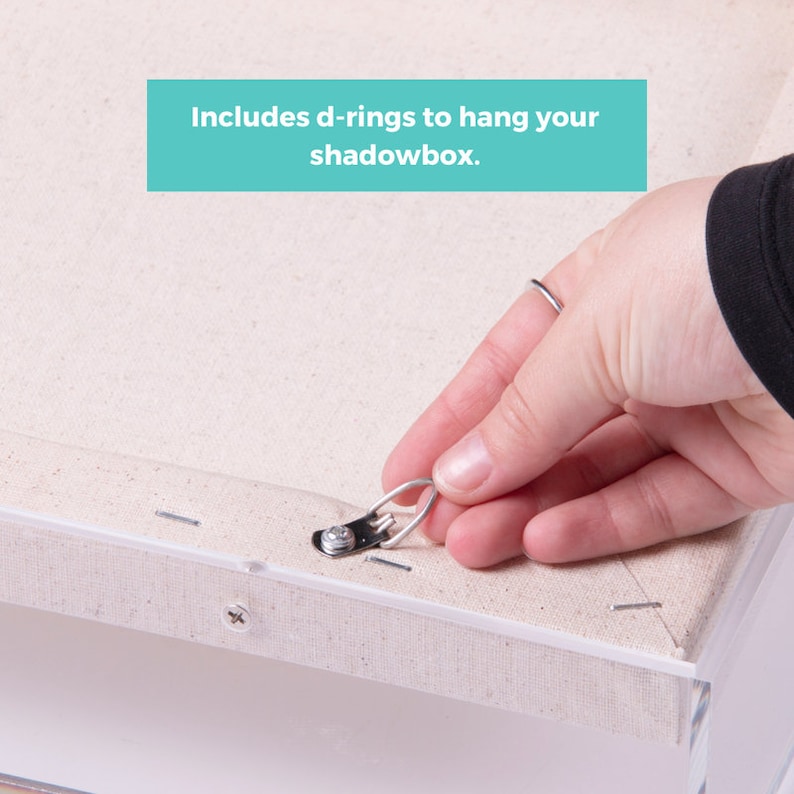 Puede incluir: Primer plano de un lienzo blanco con un anillo en D de metal sujeto a la parte posterior. El anillo en D se utiliza para colgar el lienzo en una pared. El texto "Includes d-rings to hang your shadowbox." es visible en la imagen.