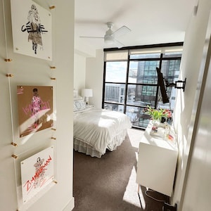 May include: A modern bedroom with a large window overlooking a city. The room features a white bed with a ruffled skirt, a white dresser, and three framed fashion illustrations hanging on the wall. A ceiling fan is visible.
