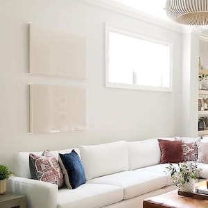 May include: A bright, airy living room with a white sofa and decorative pillows in shades of blue, burgundy, and pink. Two rectangular, framed artworks hang on the wall. A coffee table and a decorative light fixture are also visible.