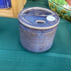 May include: A handmade ceramic container with a lid. The container is cylindrical with a dark blue and purple glaze. The lid has a small opening. The container is sitting on a green surface.