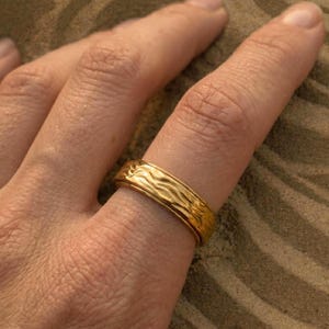 Könnte beinhalten: Ein goldfarbener Ring mit einem strukturierten, wellenartigen Muster. Der Ring wird an einem Finger getragen und zeigt sein Design und seine Handwerkskunst. Die Oberfläche des Rings reflektiert das Licht und unterstreicht seinen metallischen Glanz. Der Ring ist ein Schmuckstück.