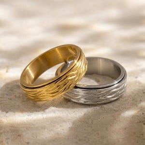 Könnte beinhalten: Zwei Ringe, einer goldfarben und einer silberfarben, werden gezeigt. Beide haben ein strukturiertes, wellenartiges Muster auf dem äußeren Band. Der goldfarbene Ring liegt vor dem silberfarbenen Ring, auf einer hellen Oberfläche.