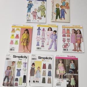May include: A collection of eight Simplicity sewing patterns for children's clothing, featuring various designs including dresses, pants, tops, and hooded outfits, displayed in their original paper envelopes with printed illustrations and model photos.