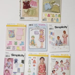 May include: A collection of eight Simplicity sewing pattern envelopes for baby clothing, featuring various designs including dresses, rompers, and hats. Each envelope displays illustrations or photographs of the finished garments, model numbers, and the Simplicity brand logo.