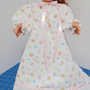 May include: A doll wearing a white nightgown with a pattern of small pink and yellow ladybugs, blue and pink flowers, and green sprigs. The gown features long sleeves with elastic cuffs, a pink ribbon tie at the neckline, and a white eyelet lace hem.