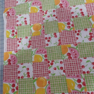 May include: Fabric with a patchwork design featuring squares of red gingham, white with cherries, green plaid, and white with fruit slices. The fruit includes oranges, watermelon, and lemons. The fabric is a light color.
