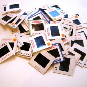 May include: A pile of old 35mm slides, each with a black frame and white border. Some slides display text such as "Kodachrome" and dates. The slides are arranged in a haphazard manner, indicating a collection of photographic memories.