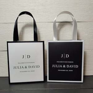 May include: Two small wedding favor bags, one white with black trim and the other black with white trim. Both have ribbon handles and the text "JD", "WELCOME TO OUR WEDDING", "JULIA & DAVID", and "NOVEMBER 22, 2025".