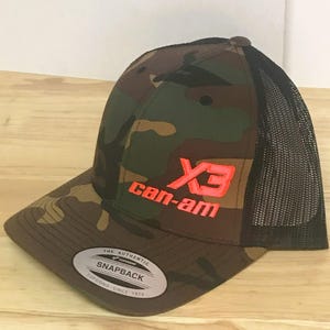 Puede incluir: Gorra de camionero de camuflaje con una parte trasera de malla negra. La gorra tiene un logotipo "X3 can-am" rojo y negro bordado en la parte delantera.