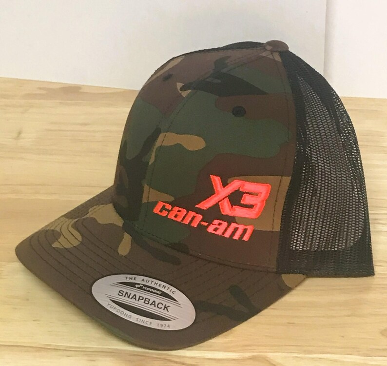 Puede incluir: Gorra de camionero de camuflaje con una parte trasera de malla negra. La gorra tiene un logotipo "X3 can-am" rojo y negro bordado en la parte delantera.