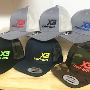 Puede incluir: Seis gorras de camionero de diferentes colores con una parte trasera de malla negra. Las gorras tienen un logotipo bordado en blanco que dice "X3 can-am" en diferentes colores.