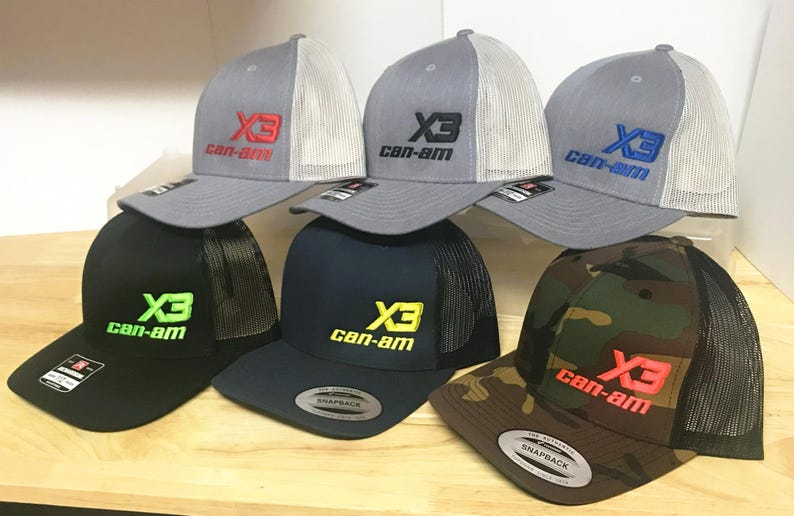 Puede incluir: Seis gorras de camionero de diferentes colores con una parte trasera de malla negra. Las gorras tienen un logotipo bordado en blanco que dice "X3 can-am" en diferentes colores.
