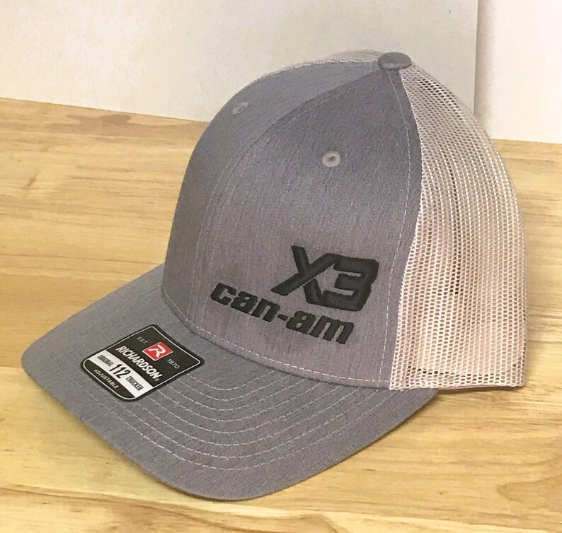 Puede incluir: Gorra de camionero gris y blanca con un logotipo bordado en negro que dice "X3 Can-Am".