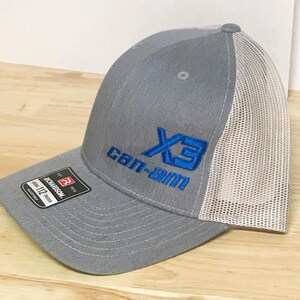 Puede incluir: Gorra de camionero de malla gris y blanca con un logotipo bordado en azul que dice "X3 Can-Am".