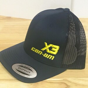 Puede incluir: Una gorra de camionero azul marino y negra con un logotipo bordado en amarillo que dice "X3 Can-Am".