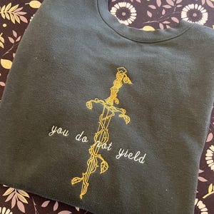 May include: Dark green sweatshirt with gold embroidered sword design and the words "you do not yield". The sword is entwined with vines and leaves. The sweatshirt is folded on a patterned surface.