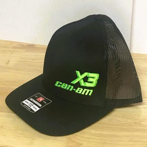 Puede incluir: Gorra de camionero negra y verde de malla con el texto "Can-Am" y un logotipo "X3" verde.