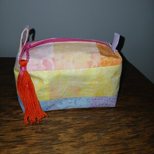 Puede incluir: Pequeño neceser rectangular de tela patchwork en colores pastel. Tiene una cremallera rosa y pequeñas trabillas de tela en los extremos. Una borla naranja cuelga del tirador de la cremallera.