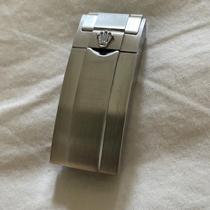 May include: A brushed stainless steel watch clasp featuring a polished silver-tone crown logo emblem at the top. The rectangular metal component has a smooth, flat surface with a central vertical line and a secure folding mechanism design.