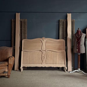 May include: A light-coloured wooden bed frame featuring a curved headboard and footboard with carved floral detailing at the centre. The piece includes two tall, vertical wooden side rails with natural wood grain textures and curved legs.
