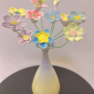 Puede incluir: Un arreglo floral de cerámica en un jarrón. El ramo presenta varias flores de colores pastel, incluyendo rosas, amarillas, azules y lavanda, con centros amarillos. El jarrón tiene forma de lágrima con un degradado de amarillo a lavanda. Las flores están sobre tallos de alambre verde.
