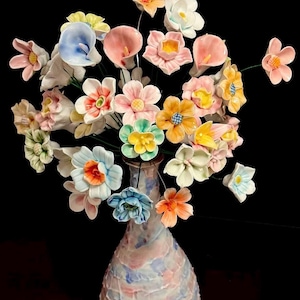 May include: A ceramic vase displaying a collection of handmade flowers in various colours. The flowers include shades of pink, yellow, orange, blue, and white. The vase has a textured, mottled surface with pink, blue, and white tones.